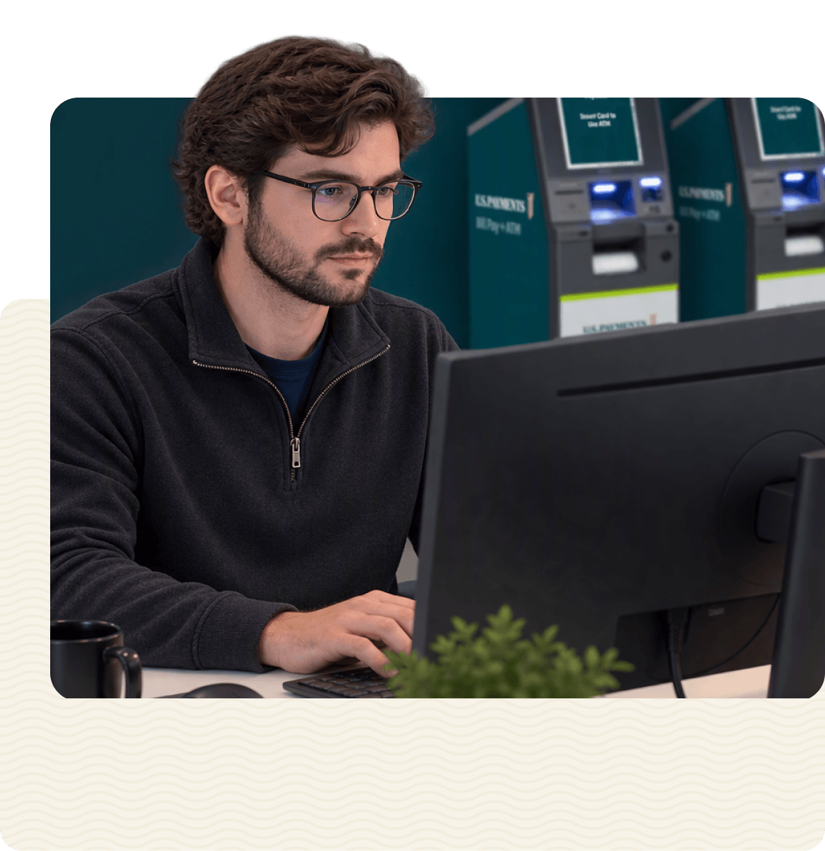 A software engineer working at a desktop monitor, with a row of bill pay and ATM kiosks lined up behind him